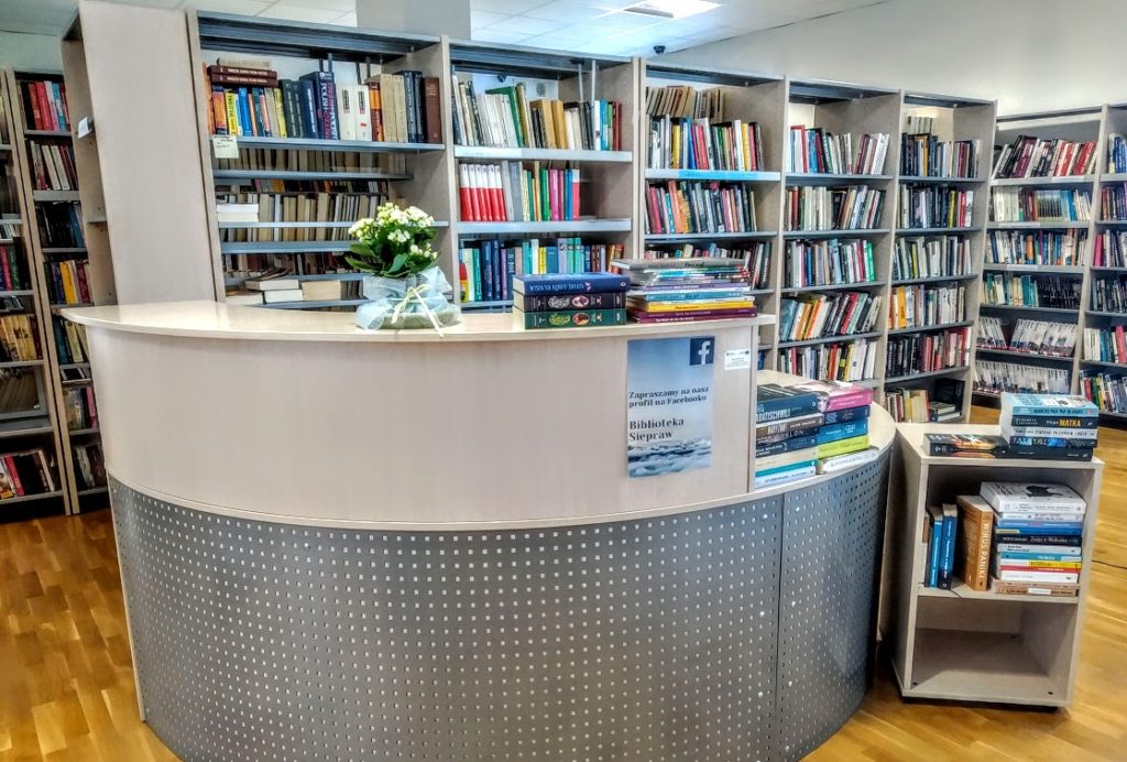 Przedstawia wnętrze biblioteki, na pierwszym planie stanowisko obsługi czytelnika